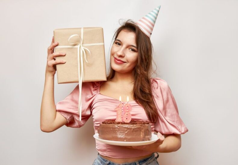 devojka drži rođendansku tortu u ruci na kojoj su svećice sa brojem 18 i poklon u ruci