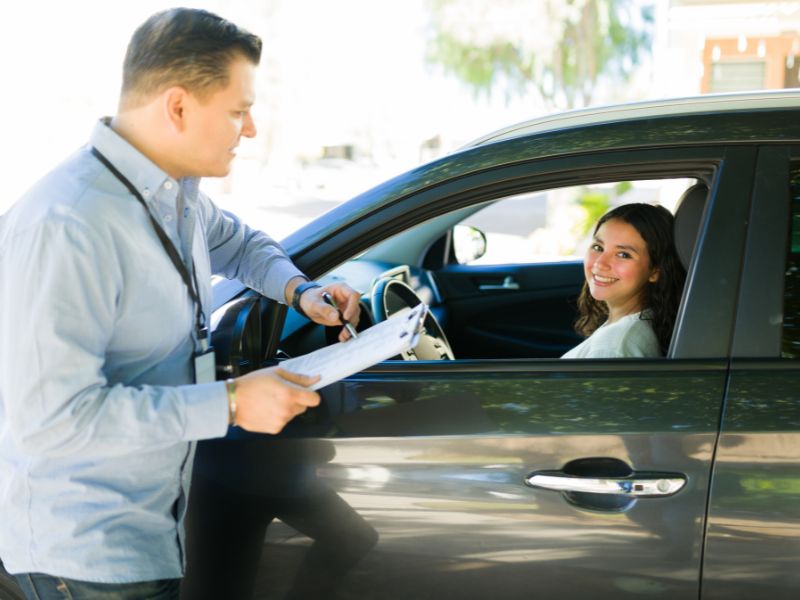 prikaz devojke koja sedi u automobilu na vozačevom mestu i instruktora sa papirima u ruci naslonjenog na automobil