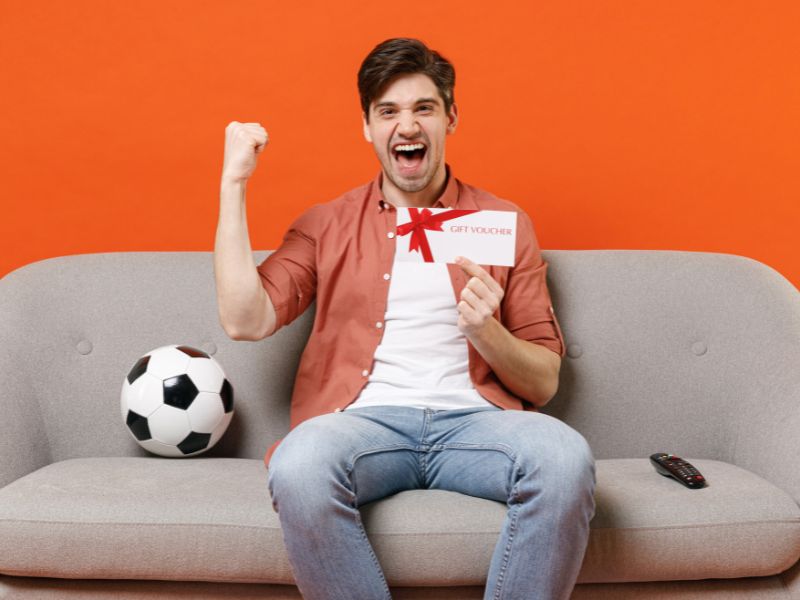 momak sedi na ugaonoj i drži sportski vaučer u ruci, sportsku loptu na ugaonoj i veselo se smeši